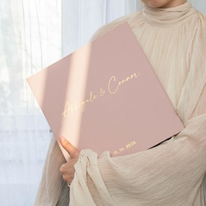 May include: A pale pink album with gold lettering reading "Abbigale & Connor" and the date "11. 14. 2025". The album is held by a person wearing a cream-coloured top. The album appears to be a wedding or event keepsake.