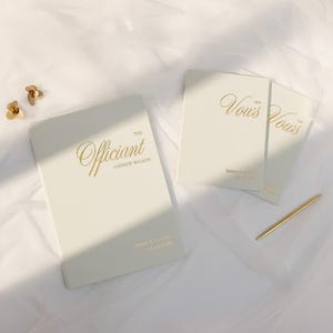 May include: Cream-coloured wedding vow books and an officiant book with gold lettering. The officiant book reads "The Officiant" and "Andrew Wilson". The vow books read "Her Vows" and "His Vows". A gold pen and decorative flowers are also present.