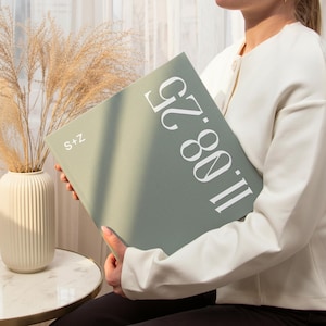 Peut inclure: Un livre vert sauge avec le texte "11.08.25" et "S+Z" en blanc. Le livre est tenu par une personne portant une veste blanche. Un vase blanc avec des plantes séchées est en arrière-plan.