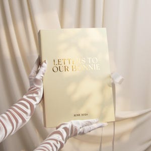 Puede incluir: Una caja de recuerdos rectangular de color crema con un cierre de cinta blanca. La portada presenta letras en lámina dorada que dicen "LETTERS TO OUR BONNIE" en la parte superior y "JUNE 2026" en la inferior, con un diseño limpio y centrado.