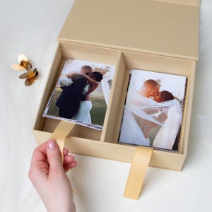 Puede incluir: Una caja de fotos beige con dos compartimentos, cada uno con fotos de boda. La caja está abierta, revelando las imágenes y una cinta dorada. Una mano tira de la cinta. Una decoración floral dorada está en la esquina superior izquierda.