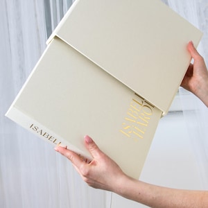 May include: A cream-colored box with a matching lid is held open, revealing a book. The book and box feature gold lettering that reads "ISABELLA HAROT". The box is held by two hands, and the background is a soft white.