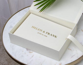 May include: An open, off-white box with a matching lid, revealing a cream-colored book inside. The book cover is embossed with "HELEN & FRANK" and "JUNE 20, 2026" in gold lettering. A white ribbon is attached to the box.