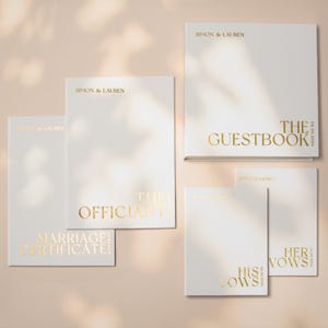 Puede incluir: Un conjunto de cinco libros blancos con letras en lámina de oro. Las portadas presentan los nombres Simon & Lauren, la fecha 20.08.2026 y los títulos The Guestbook, The Officiant, Marriage Certificate, His Vows y Her Vows en fuente serif.