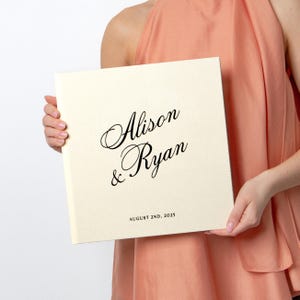 May include: A cream-coloured wedding album with the names "Alison & Ryan" in elegant black script. The date "AUGUST 2ND, 2025" is printed below. The album is held by a person wearing a peach-coloured top.