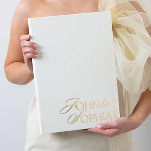 May include: A cream-colored wedding album with gold lettering that reads "John & Sophia" and the date "08.25.2025". The album is held by a person.