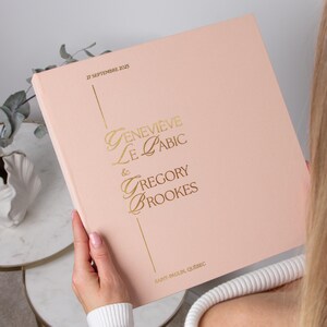 May include: A pale pink wedding album with gold lettering. The cover displays the names "Genevieve Le Cabic & Gregory Brookes" and the date "27 Septembre 2025". The location "Saint-Paulin, Quebec" is printed at the bottom.