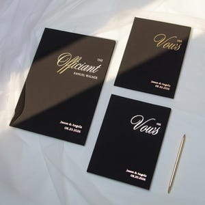 Może przedstawiać: Trzy czarne księgi ślubnych przysięg z złotymi i białymi literami. Jedna księga głosi "The Officiant", a druga "Her Vows". Trzecia księga głosi "His Vows". Obecny jest również złoty długopis.