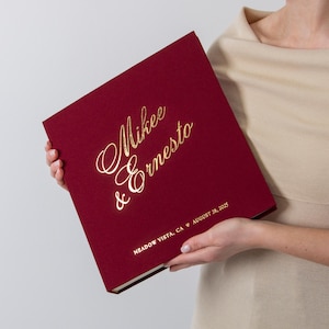 May include: A burgundy photo album with gold lettering that reads "Mikee & Ernesto" and the date August 28, 2025. The album is held open, showcasing its cover design. The album is a wedding keepsake.