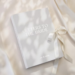 Puede incluir: Un libro de tapa dura gris claro titulado LETTERS TO THE BRIDE con la fecha 19.09.2026 impresa en la portada. El libro presenta una cinta de satén blanca atada en el lateral y se muestra sobre un fondo de tela suave de color blanco roto.