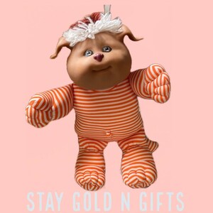May include: A plush toy dog with a white and orange striped outfit. The toy has a brown face with a pink nose and blue eyes. The toy is wearing a white and red hat with a white pom-pom.