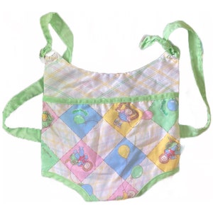 May include: A vintage Cabbage Patch Kids baby carrier. The carrier features a patchwork design with pastel squares and Cabbage Patch Kids graphics. It has green straps and trim, and is made of fabric.