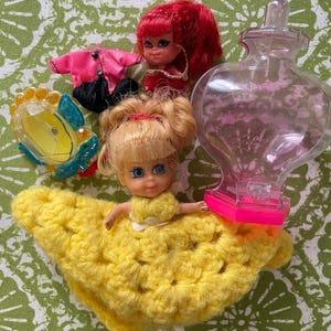 May include: A vintage doll with blonde hair and blue eyes wearing a yellow crocheted dress. The doll is holding a small, clear plastic bottle with a pink lid. There is also a pink shirt, black pants, and a clear plastic mirror with a blue frame.