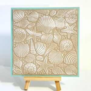 May include: A square, tan-colored artwork featuring an embossed design of various seashells and starfish. The artwork is framed with a light teal border and displayed on a small wooden easel. The design includes detailed textures and shapes of different marine life.