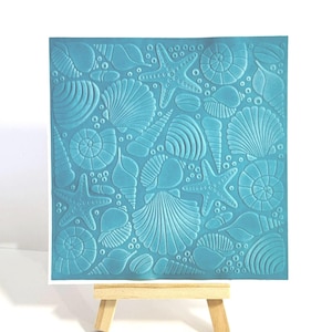 May include: A square, turquoise-colored art piece featuring an embossed design of various seashells and starfish. The artwork is displayed on a small wooden easel, showcasing the intricate details of the marine-themed relief.
