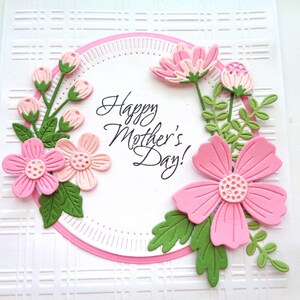 May include: A white card with a pink and green floral design. The card has a circle in the center with the text "Happy Mother's Day!" in black cursive writing.
