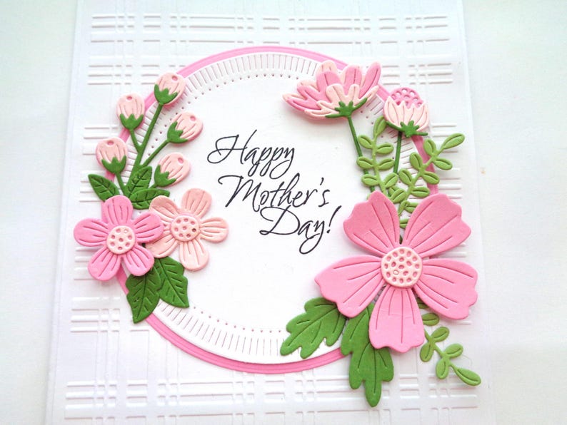 May include: A white card with a pink and green floral design. The card has a circle in the center with the text "Happy Mother's Day!" in black cursive writing.
