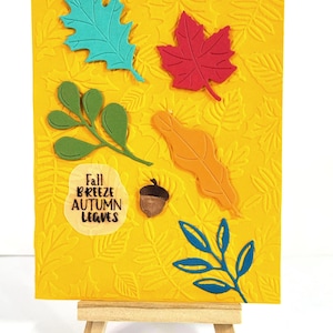 May include: A rectangular yellow card featuring an embossed leaf pattern, decorated with a teal oak leaf, a red maple leaf, a green sprig, an orange leaf, a small brown acorn, and a blue sprig. The text "Fall BREEZE AUTUMN LEAVES" is printed.