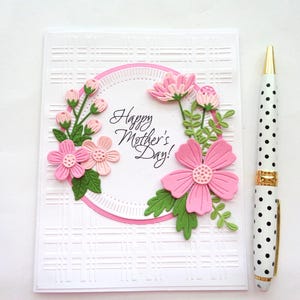 May include: A white card with a pink and green floral design and the text "Happy Mother's Day!" in a circle. The card has a white and black polka dot pen next to it.