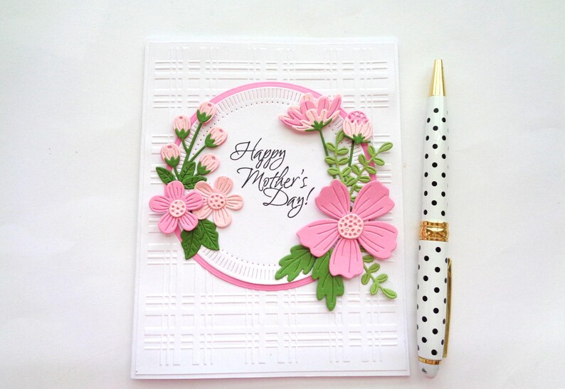May include: A white card with a pink and green floral design and the text "Happy Mother's Day!" in a circle. The card has a white and black polka dot pen next to it.