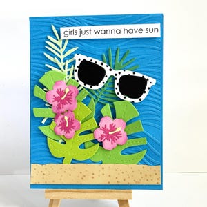 May include: A handmade greeting card with a tropical theme. It features a blue background with a wave pattern, green leaves, pink flowers, and white sunglasses with black polka dots. The text "girls just wanna have sun" is displayed.