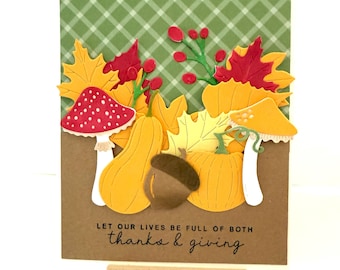 May include: A handmade greeting card with a fall theme. The card features paper cutouts of yellow and red leaves, pumpkins, mushrooms, and an acorn. The background is green plaid. The text reads, "Let our lives be full of both thanks & giving."