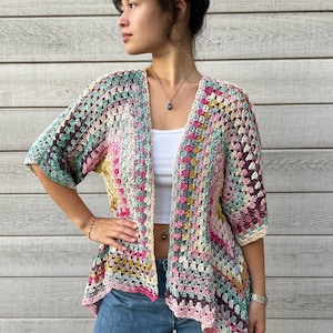 Op de afbeelding: Een kleurrijk, opengebreid vest in een granny square patroon. Het vest heeft een mix van roze, groene, gele en paarse tinten. Het kledingstuk met korte mouwen wordt gedragen over een witte top en een blauwe spijkerbroek.