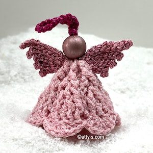 Op de afbeelding: Een roze gehaakt engel ornament met een roze en witte glitter afwerking. De engel heeft vleugels, een jurk en een roze kralenhoofd met een roze garen halo.
