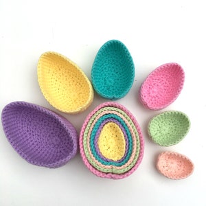 Op de afbeelding: Een set van zeven gehaakte, eivormige mandjes in verschillende pastelkleuren, waaronder paars, geel, groen, roze en blauw. De mandjes zijn in elkaar genest, waardoor een regenboog-effect ontstaat.