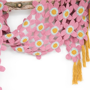 Op de afbeelding: Een roze gehaakte sjaal met bloemmotieven en gele kwastjes. De bloemen hebben een lichtblauwe kern en een gele kern.