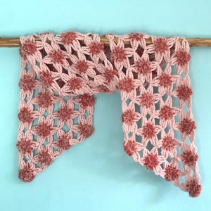 Può includere: Una sciarpa all'uncinetto rosa e rossa con un motivo a fiore e pompon. La sciarpa è appesa a un bastone di legno.