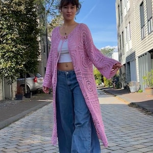 Op de afbeelding: Een lange, open roze gehaakte cardigan met klokmouwen. De cardigan heeft een gedetailleerd cirkelvormig patroon en wordt gedragen over een witte top en een blauwe jeans. De outfit is gestyled voor een casual, bohemien look.
