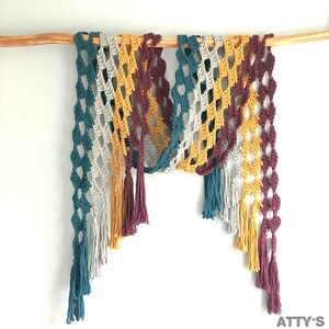 Op de afbeelding: Een kleurrijke, open geweven gehaakte sjaal met ruitvormige patronen. De sjaal heeft kwastjes in verschillende kleuren, waaronder blauwgroen, grijs, mosterdgeel en bordeauxrood. Het wordt getoond op een houten stok tegen een witte achtergrond.