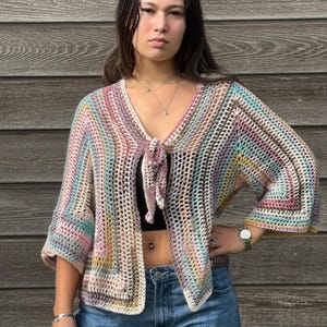Op de afbeelding: Een veelkleurig gehaakt topje met een strik aan de voorkant en wijde mouwen. Het topje wordt gedragen over een zwart tanktopje en een blauwe jeans.