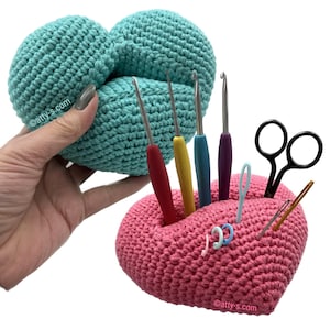 May include: A pink crocheted heart-shaped holder with a variety of crochet hooks and scissors inside. The holder is sitting on a white surface. A turquoise crocheted heart is in the background.