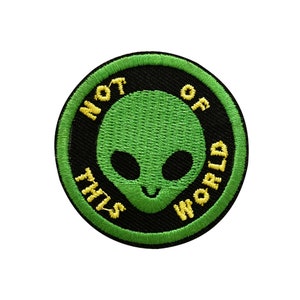 May include: Green embroidered patch with a black background and yellow text that reads "Not of this world". The patch features a green alien with black eyes and a smile.