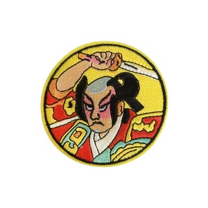 May include: A round embroidered patch featuring a cartoon samurai warrior with a sword, wearing a red and yellow robe. The background is yellow.