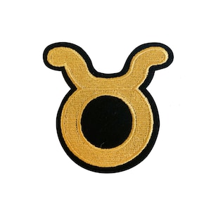 May include: Gold embroidered Taurus zodiac sign patch. The patch is outlined in black and has a black circle in the center.