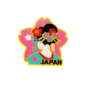 May include: Embroidered patch featuring a woman in a traditional Japanese kimono with a cherry blossom design. The patch is pink, blue, yellow, and white with the word "JAPAN" in black lettering.