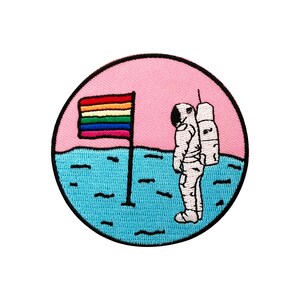 May include: A round embroidered patch with a pink background, a blue and white astronaut standing on a blue surface with a rainbow flag planted in the ground.
