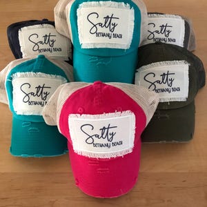 May include: Five trucker hats with a distressed look and a white patch with the text "Satty Bethany Beach" in black script. The hats are in various colors including teal, pink, black, and green.