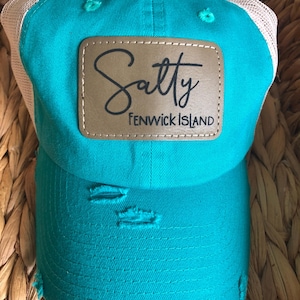 May include: A turquoise blue and white mesh trucker hat with a distressed look. The hat has a brown leather patch with the text "Salty Fenwick Island" in black.