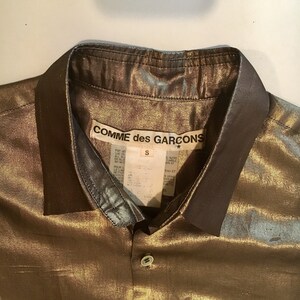 K&ouml;nnte beinhalten: Ein goldfarbenes, metallisches Hemd mit Kn&ouml;pfen und schwarzem Kragen. Das Hemd hat ein Etikett mit der Aufschrift "COMME des GARCONS" und ein Gr&ouml;&szlig;enetikett mit der Aufschrift "S".