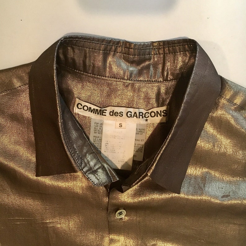 K&ouml;nnte beinhalten: Ein goldfarbenes, metallisches Hemd mit Kn&ouml;pfen und schwarzem Kragen. Das Hemd hat ein Etikett mit der Aufschrift "COMME des GARCONS" und ein Gr&ouml;&szlig;enetikett mit der Aufschrift "S".