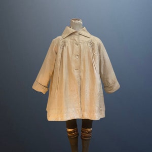 May include: A beige, long-sleeved coat with a button closure and a small collar. The coat has a gathered detail at the shoulders and a slightly flared silhouette.