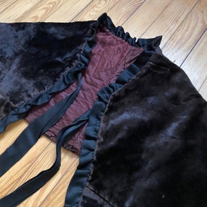 以下が含まれることがあります： 黒のベルベットのケープで、フリルトリムと黒いサテンのリボンが付いています。ケープはワインレッドの生地で裏打ちされています。