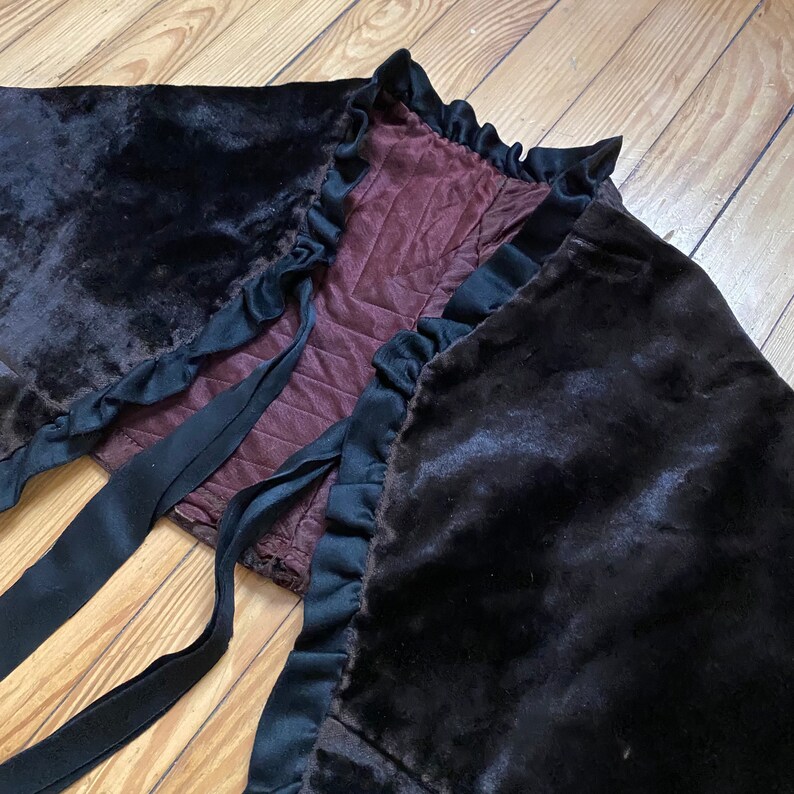 以下が含まれることがあります： 黒のベルベットのケープで、フリルトリムと黒いサテンのリボンが付いています。ケープはワインレッドの生地で裏打ちされています。