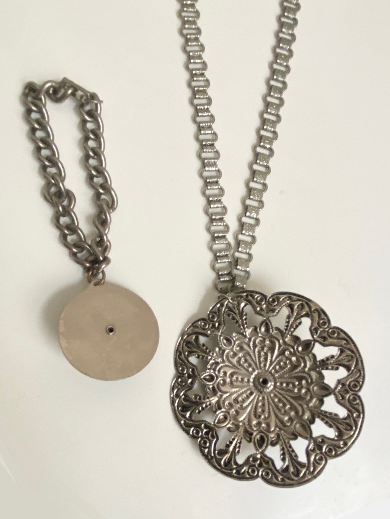 K&ouml;nnte beinhalten: Eine silberne Kettenkette mit einem gro&szlig;en, verzierten, runden Anh&auml;nger. Der Anh&auml;nger hat ein filigranes Design und ist mit einem kleinen, runden Verbinder an der Kette befestigt.