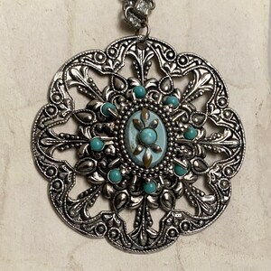 K&ouml;nnte beinhalten: Eine silberne Halskette mit einem gro&szlig;en, verzierten Anh&auml;nger, der einen t&uuml;rkisfarbenen Stein zeigt, der von kleineren t&uuml;rkisfarbenen Steinen umgeben ist. Der Anh&auml;nger hat die Form einer Blume mit filigranen Details.