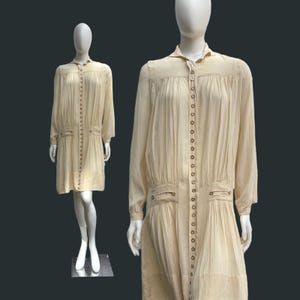 Könnte beinhalten: Ein cremefarbenes Vintage-Kleid mit langen Ärmeln und Knopfleiste vorne. Das Kleid hat Falten an Schultern und Taille sowie kleine Taschen. Das Kleid wird auf einer Schaufensterpuppe präsentiert.